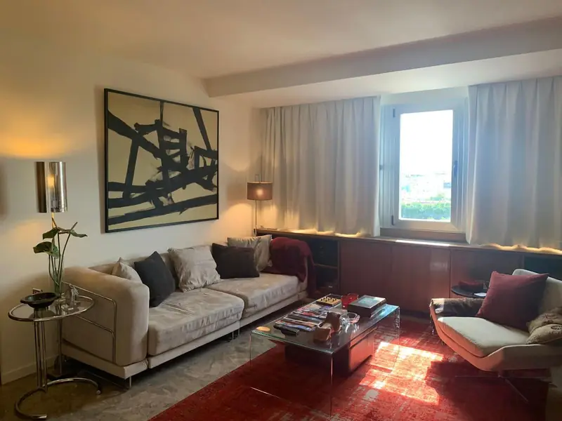 Sala com tapete carmim, sofá claro, pintura abstrata negra e luz rasante