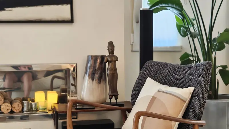 Cadeirão de tweed e madeira junto a um nicho de lenha com velas, figura em bronze e vaso de pele