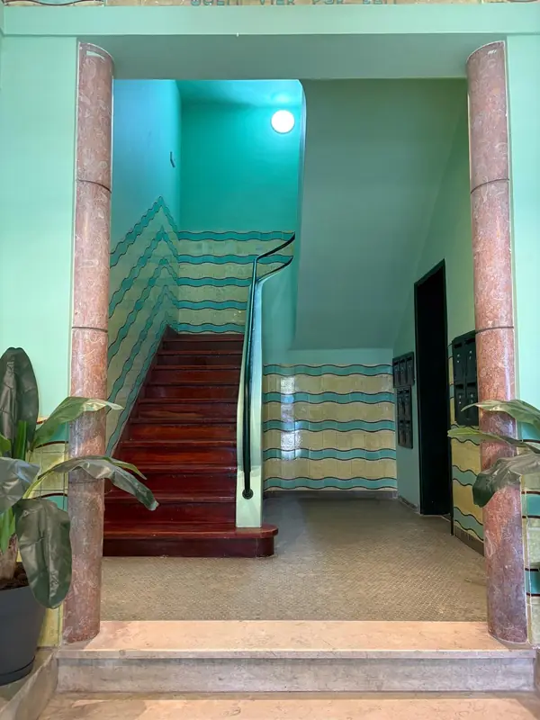 Escada do prédio com azulejo verde-água, degraus de madeira e colunas de mármore rosa
