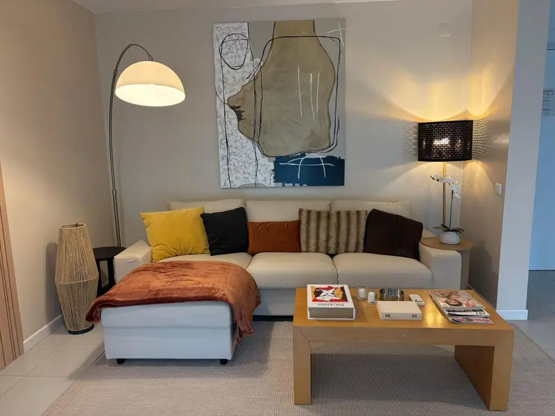 Sala com sofá de canto claro, manta cor de ferrugem, candeeiro de arco e retrato abstrato