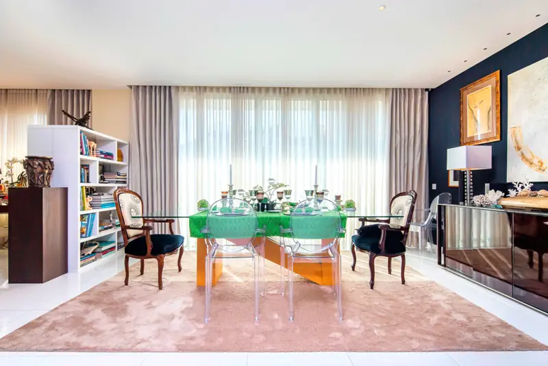 Sala de jantar com mesa de vidro verde, cadeiras estofadas, parede negra e estante branca
