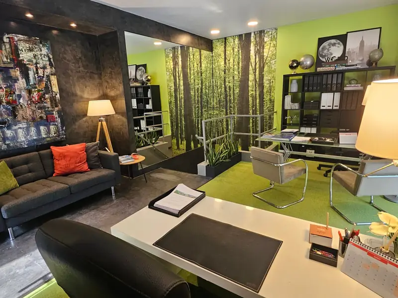 Escritório com papel de parede de floresta, alcatifa verde e sofá cinzento com almofada coral