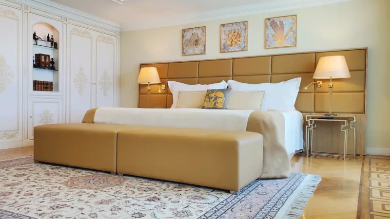 Quarto com cabeceira estofada dourada, banco aos pés da cama e tapete persa