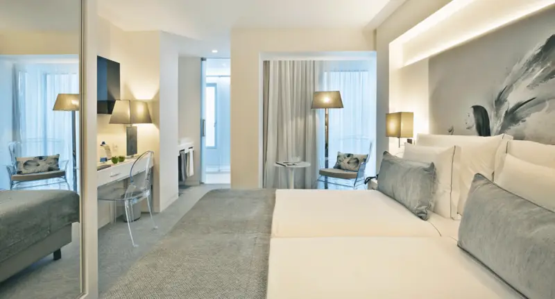 Quarto de duas camas com almofadas cinzentas, candeeiros de pé e mural a preto e branco