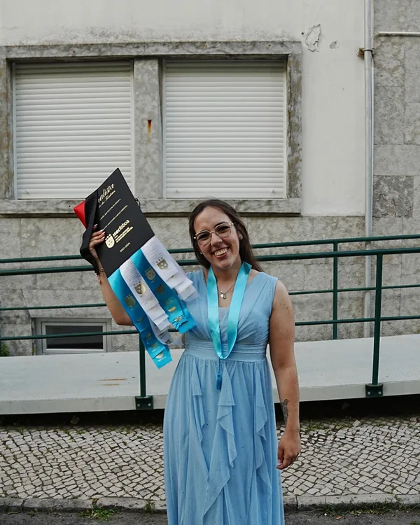 Beatriz sorridente, de vestido azul, com o diploma e as fitas da licenciatura.